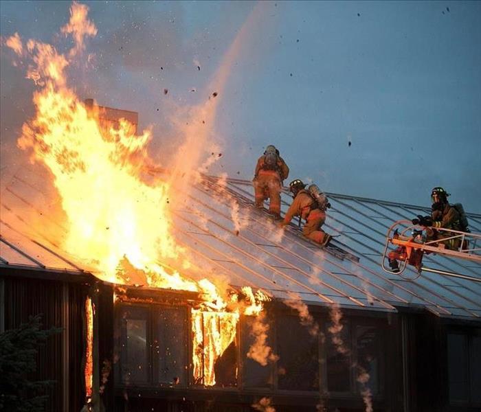 3 firefighters on top of the roof of a home engulfed in flames.