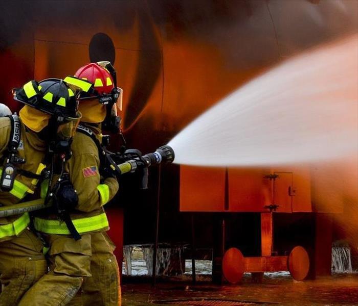 Two firefighters with a fire hose fighting against a large fire.
